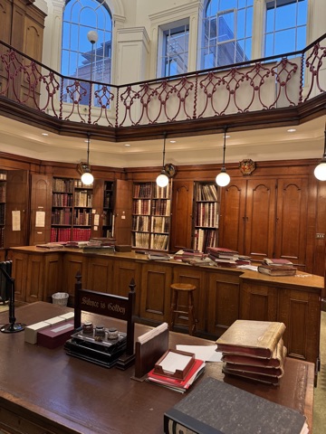 A panelled library under a gallery with a red iron balustrade and arched windows: shelves of bound volumes behind open cupboard doors, ledgers stacked on the counter, and a desk in front with a notice reading Silence is Golden.
