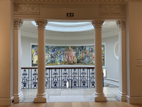 A pair of white columns framing a curved landing under an oval skylight, a blue iron balustrade in front, and a wide, loosely painted canvas of figures in blues, yellows and pinks on the far wall.
