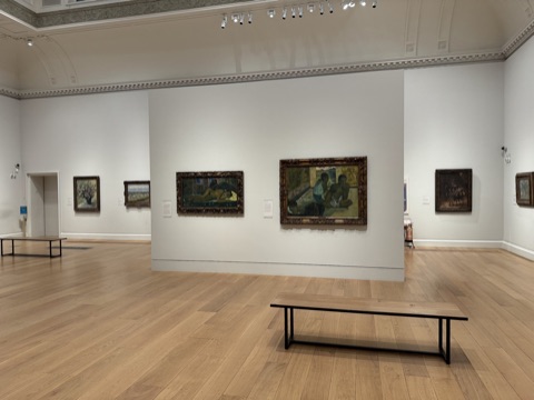 A high white gallery with a moulded ceiling and a pale wood floor: two Gauguins in carved gold frames on a free-standing wall, more paintings on the walls around, one plain bench and nobody in the room.