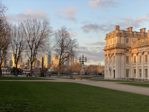 A lawn at the Old Royal Naval College in low sun, one classical range lit gold and Canary Wharf behind the trees