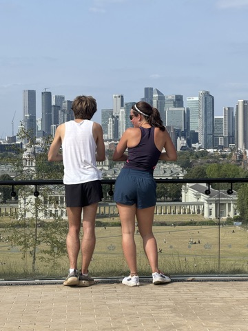 Two people at the railing on Greenwich Hill, looking across the park to Canary Wharf's towers