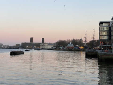 Greenwich across the water at dusk, a green dome by the river and the Cutty Sark's masts against a pink sky