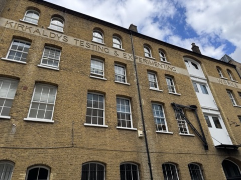 The front of Kirkaldy's Testing Works on Southwark Street, seen from the pavement: four storeys of yellow London brick with arched windows, the words Kirkaldy's Testing & Experimenting Works cut into a stone band across the second floor, white loading doors and an iron hoist on the right, under a blue sky with white clouds.