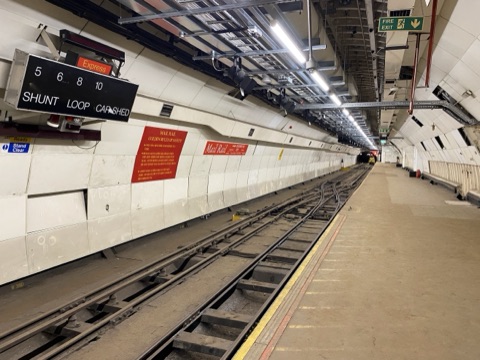 A Mail Rail platform: narrow track and points running away down a white-panelled tunnel, an indicator board overhead