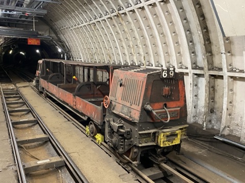 A rusty red battery locomotive coupled to open wagons, standing on narrow track inside a bolted cast-iron tunnel