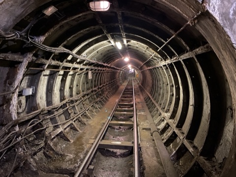 A narrow railway tunnel of riveted iron rings, a single track running away towards distant lights