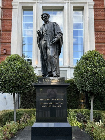 A bronze statue of Michael Faraday in a long gown, one hand raised, on a black granite plinth lettered in gold with his name and dates, between two clipped bay trees in front of a red-brick and stone facade with tall windows.