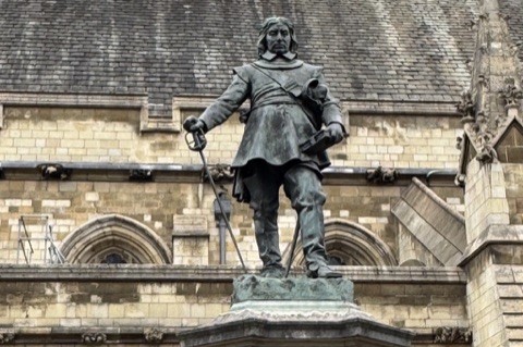 Hamo Thornycroft's bronze statue of Oliver Cromwell on its stone plinth, a sword point-down in one hand and a book in the other, with the Gothic stone wall and slate roof of the Palace of Westminster behind