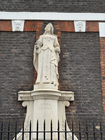 A white stone statue of Queen Anne in a long embroidered gown and lace collar, standing in a shallow niche in the brick wall on a scrolled pedestal lettered ANNA REGINA, a carved stone head set in the brickwork either side of her, and iron railings across the foot.