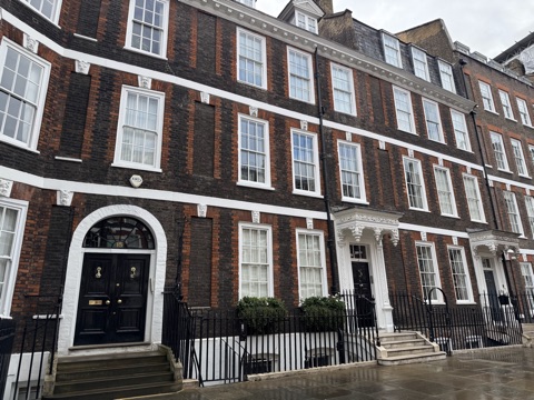 A terrace of early Georgian houses in dark brown brick with red brick dressings and white-painted bands: tall white sash windows, a carved white stone head as the keystone over each window opening including the blind ones, deep carved door hoods over black front doors, and number 15 with a fanlight, behind black iron railings on a wet pavement.