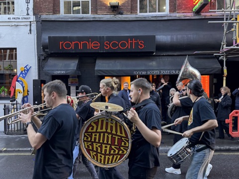 A brass band in black T-shirts marching past the club's black frontage on Frith Street, its name in red neon above the awnings: a trumpet and a saxophone in front, a bass drum reading Soho Saints Brass Band, a snare and a sousaphone behind, and people watching from the doorway.