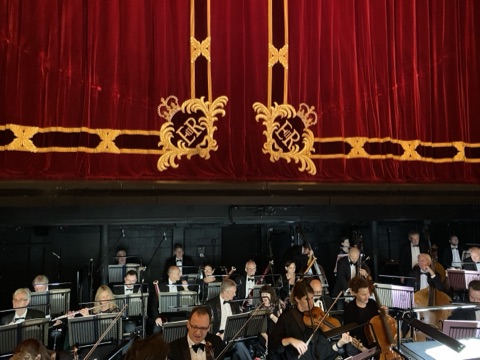 The orchestra pit from the stalls before a performance: players in black tie at their stands with flutes, bassoons, violins, a cello and double basses, under the red velvet house curtain with its gold braid and crowned EIIR ciphers.