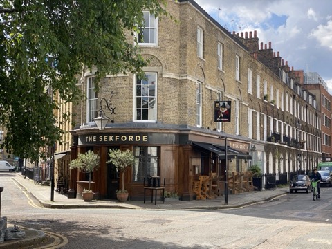 The Sekforde on its corner: a yellow-brick Georgian terrace curving round the junction, a dark pub front with the name in gold, olive trees in pots by the door and a wrought-iron lamp bracket above.