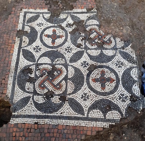 A square mosaic panel of black, white and red pieces with two knot patterns and roundels of four-petalled flowers, set in a surround of red tiles