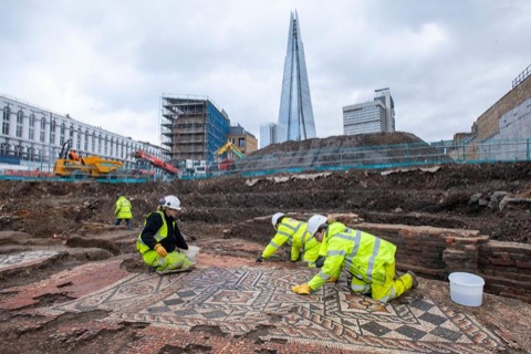 Four archaeologists in high-visibility jackets and hard hats kneeling on a large Roman mosaic of black, white and red pieces in an open excavation, with the Shard and cranes behind them
