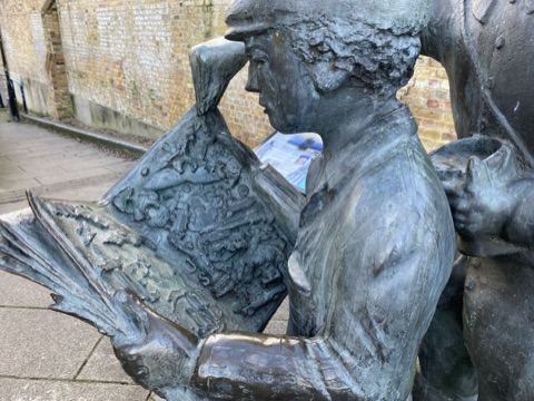 A bronze pilgrim reading a comic over the shoulder of a boy in a flat cap, a dog up on its hind legs beside them