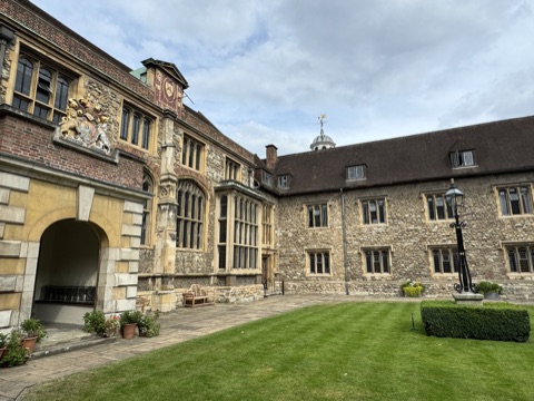 A grass court inside the Charterhouse, the hall range in flint and stone with a carved coat of arms and a red sundial