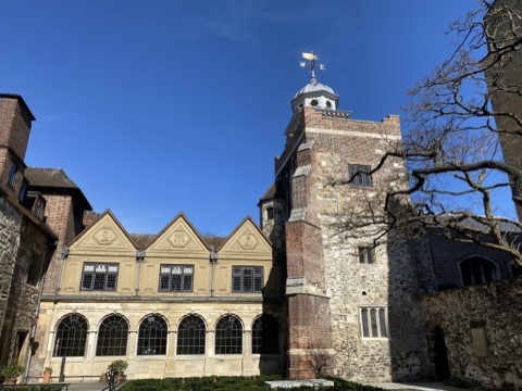 A brick and flint tower with a cupola and gilded weathervane, above an arcaded cloister of three gables