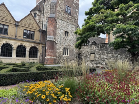 A garden of clipped box in a knot pattern and a bed of yellow rudbeckias, purple geraniums and red spikes of flowers, in front of a stone and brick tower with leaded windows, a gabled Tudor range on the left and an old flint wall on the right.