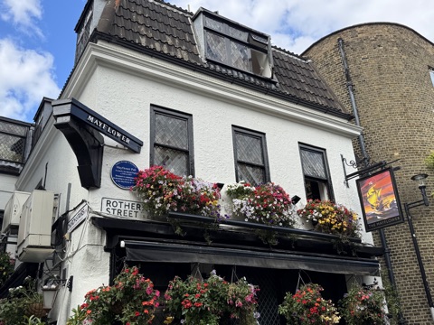 The Mayflower's white-rendered corner seen from the pavement: a black MAYFLOWER board on a bracket, leaded diamond-pane windows, window boxes and hanging baskets in full flower along the first floor, and a swinging sign painted with a galleon under a red sky.