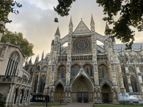 The abbey's north front at dusk: a great rose window above three deep pointed porches crowded with carved figures, pinnacled turrets either side, and plane trees in leaf overhanging the view.