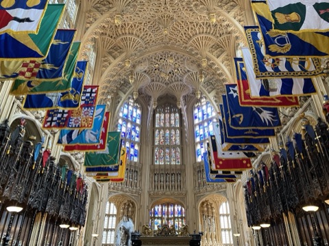 The fan vault of the Lady Chapel from below: carved stone fanning out into hanging pendants across the whole ceiling, rows of heraldic banners in green, blue, red and gold along both sides above the stalls, and tall stained glass at the east end.