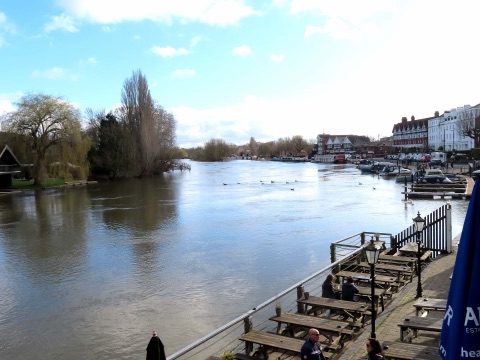 The Thames at Henley in spring, wide and full, with willows on the far bank, boats moored by the town's riverside houses and pub tables on the bank in front.