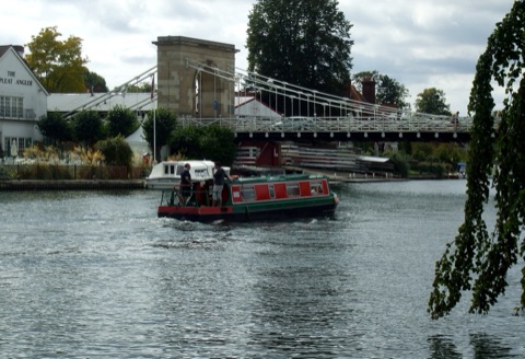 A green-and-red narrowboat passing under Marlow's white suspension bridge on the Thames, with a riverside pub on the left bank and trees beyond.