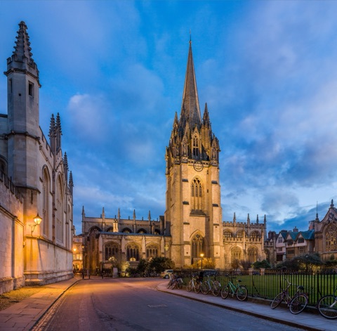 The spire of the University Church of St Mary in Oxford lit at dusk, with a gas lamp on the wall of All Souls to the left and bicycles against the railings.
