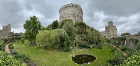 Windsor Castle's grey stone towers above the moat garden in summer, with lawns, flower beds and clipped shrubs on the slope below the walls.