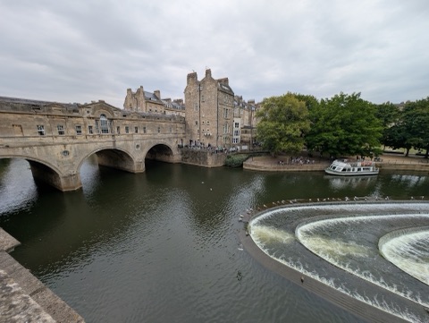Pulteney Bridge's arches and the shops built along it above a horseshoe weir, with a pleasure boat on the river and trees on the bank.