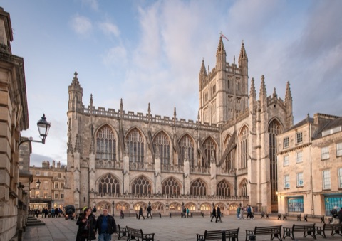 Bath Abbey's west front and tower of pale stone with tall pinnacles, in evening light, with people crossing the paved square in front.