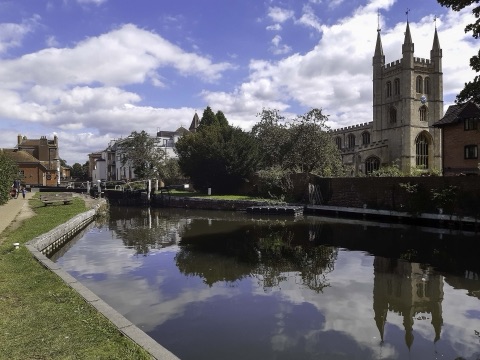 A canal lock with its gates and a wide pound of still water reflecting the sky, a church tower with pinnacles on the bank behind, and a towpath on the left.