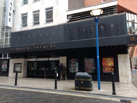 The Peacock Theatre's black frontage on Portugal Street: its name in gold letters over a row of dark glass doors, gilt starbursts along the fascia above, LSE on the canopy to the right, and lit poster cases beside the entrance.