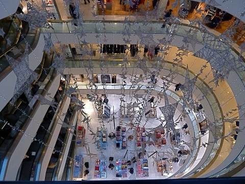 Looking down the shop's oval atrium from the top floor: escalators and galleries ringing the well, cosmetics counters on the floor far below, and a chandelier of wire stars hung down the middle.