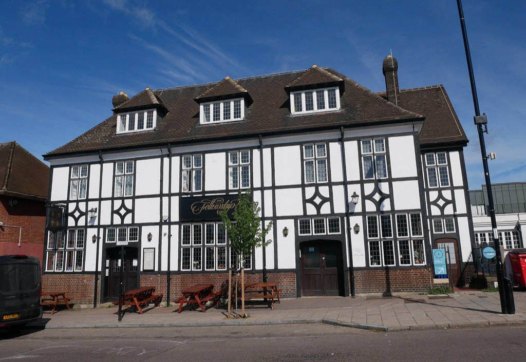A large black-and-white timbered pub filling a street corner under a steep tiled roof, benches on the pavement in front of it, under a clear blue sky.