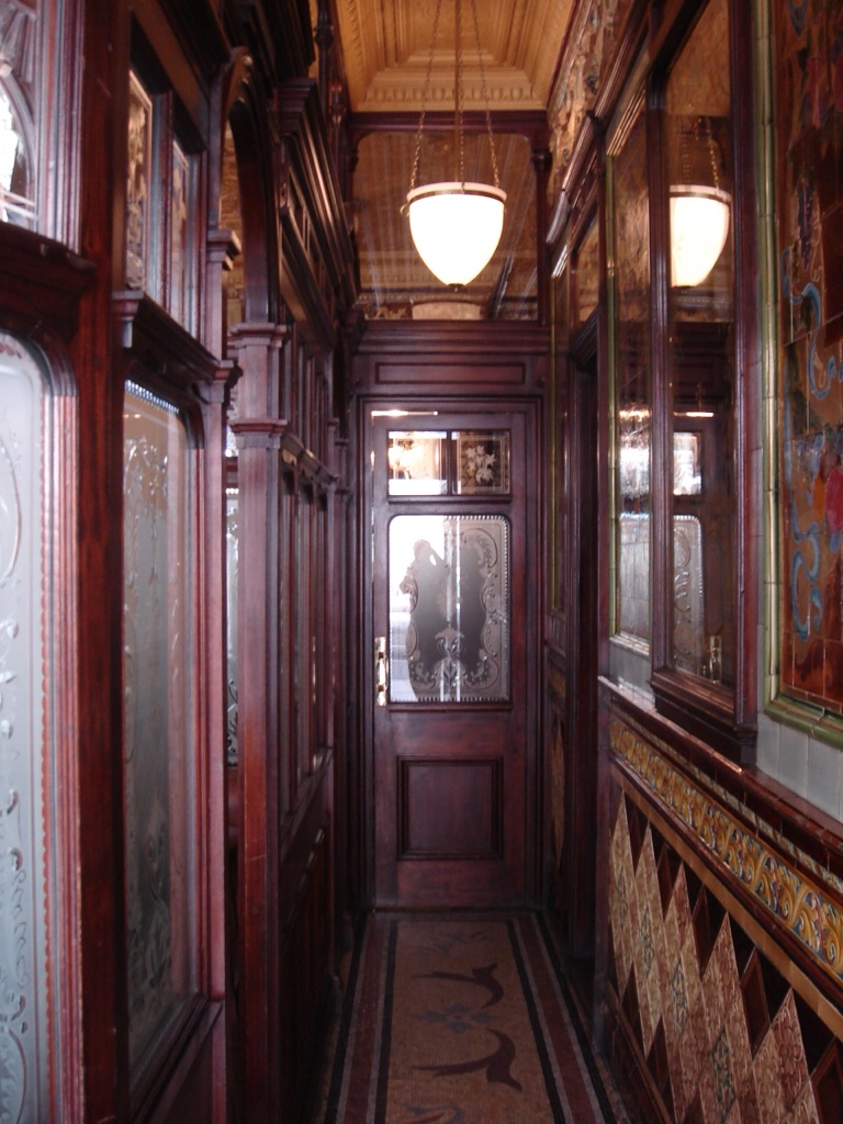 A narrow corridor inside a Victorian pub, mahogany screens set with etched glass down both sides, polychrome tiles below and a gilded ceiling above, lit by two globe lamps.