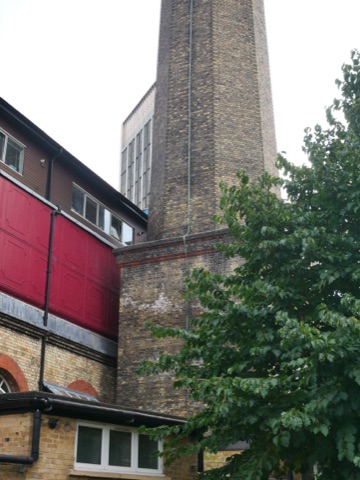 The base of the station's tall brick chimney beside a red-painted loading door and a modern glass block behind, with a tree in leaf in front.