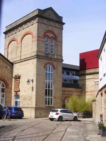 The pumping station's tall square tower of yellow brick with red-brick arches, a round-headed window up its face and a stone tablet reading The London Hydraulic Power Co 1902, seen across a cobbled yard with old rail tracks and two parked cars.