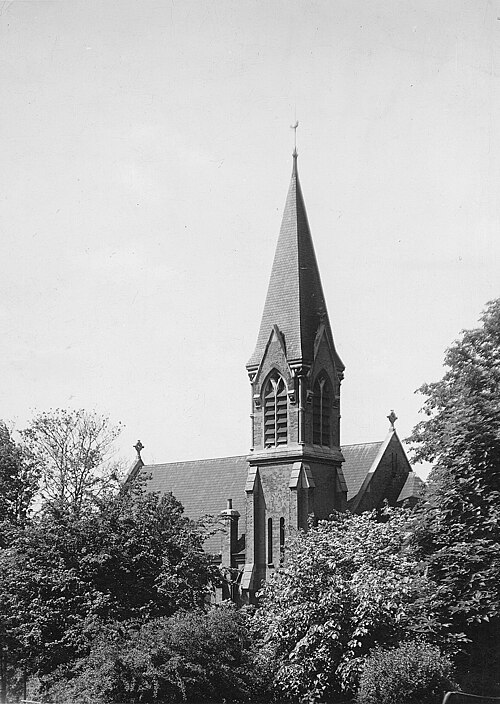 A black-and-white photograph of a Victorian Gothic church: a square brick tower carrying a tall slate broach spire, lancet belfry openings under it, the nave roof running back behind, and trees crowding in below.