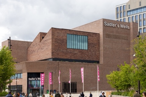 A new theatre of dark red brick in stepped blocks, the name Sadler's Wells in white letters high on its blank flank, a wide picture window set into the middle block, pink banners on poles by the glass entrance and people crossing the square in front.
