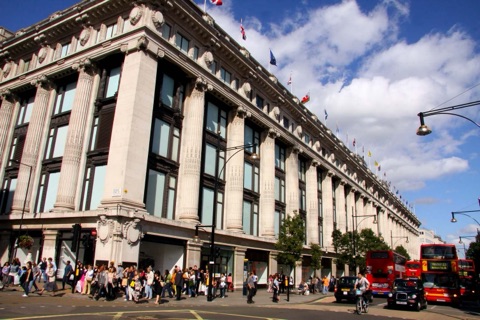 Selfridges' long stone front on Oxford Street: giant Ionic columns rising through three storeys of windows, flags along the parapet, and buses, taxis and shoppers on the street below.