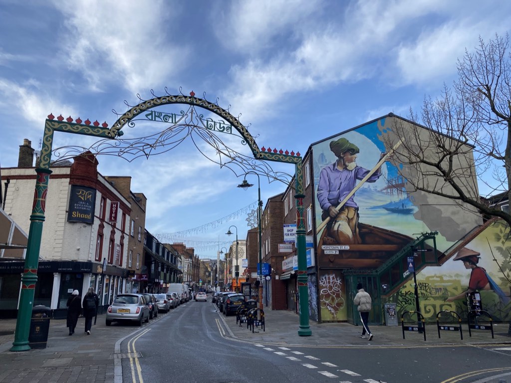 A green and gold arch lettered Bangla Town spanning Brick Lane, a mural of a boatman on the gable beside it