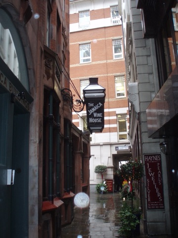 Looking down the wet alley between dark stone buildings: a large hanging lantern lettered The Jamaica Wine House out over the passage, JAMAICA BUILDINGS carved on the left, a jeweller's red sign on the right, and the street sign at the far end where the alley runs under a building.