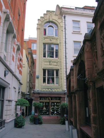 A very narrow decorated stone shopfront wedged between taller buildings where two alleys meet: an arched window at the top and two sash windows below it in moulded surrounds, a lit jeweller's window at street level under the name John Haynes Jewellers, bay trees and pots of geraniums on the flagstones, and a hanging sign for the George and Vulture on the wall to the left.