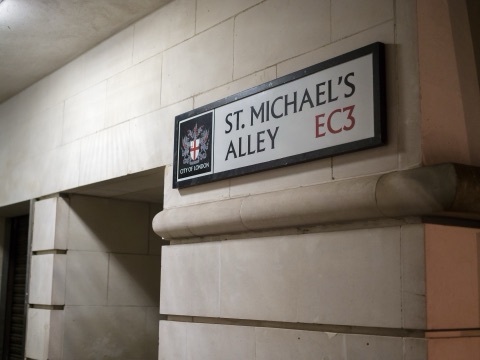 A City of London street sign reading ST. MICHAEL'S ALLEY EC3, the City's arms in a panel beside the lettering, screwed to a Portland stone wall above a moulded course, lit at night.