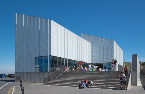 The Turner Contemporary's white ridged gallery blocks beside the sea on a bright day, with people on the steps and deckchairs on its terrace.