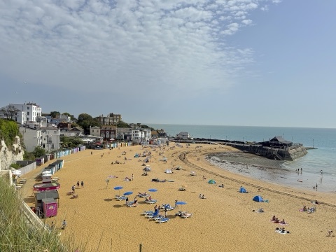 Broadstairs' curved sandy bay full of parasols and sunbathers, with houses on the cliff, a stone jetty and the sea beyond under high cloud.