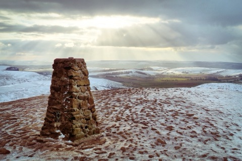 A stone trig point on a hilltop in snow, with shafts of sun breaking through cloud over a white valley and hills beyond.