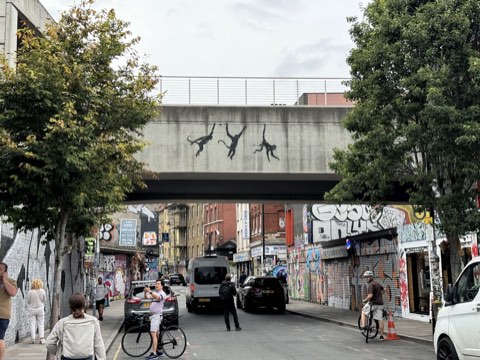 Three black stencilled monkeys by Banksy swinging across the concrete railway bridge over Brick Lane, graffiti-covered shopfronts beneath